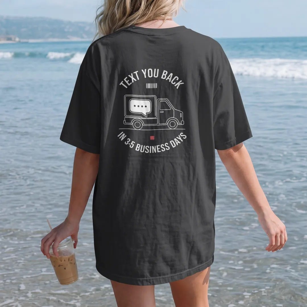 Woman on the beach in a Black tee, back view: minimal single-line drawing of a small delivery truck carrying a chat bubble, a tiny barcode beside it, with "TEXT YOU BACK / IN 3-5 BUSINESS DAYS" in small spaced-out caps, printed in bone white with a…