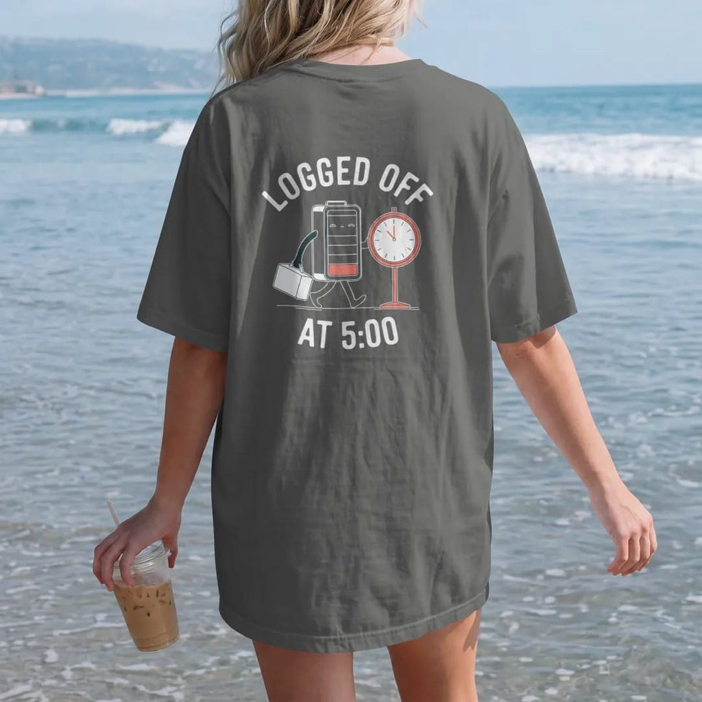 Woman on the beach in a Pepper tee, back view: minimal single-line drawing of a sleepy battery walking away from a punch clock carrying a lunchbox, with "LOGGED OFF / AT 5:00" in small spaced-out caps, printed in bone white with a low-battery red…