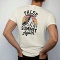 Man wearing a Ivory tee, seen from the back: a tired cartoon hiker cresting a ridge to see a taller peak behind it, stacked text above, faded 70s souvenir style, lettered "FALSE SUMMIT / AGAIN" in chunky rounded 70s letters with a drop shadow and a…