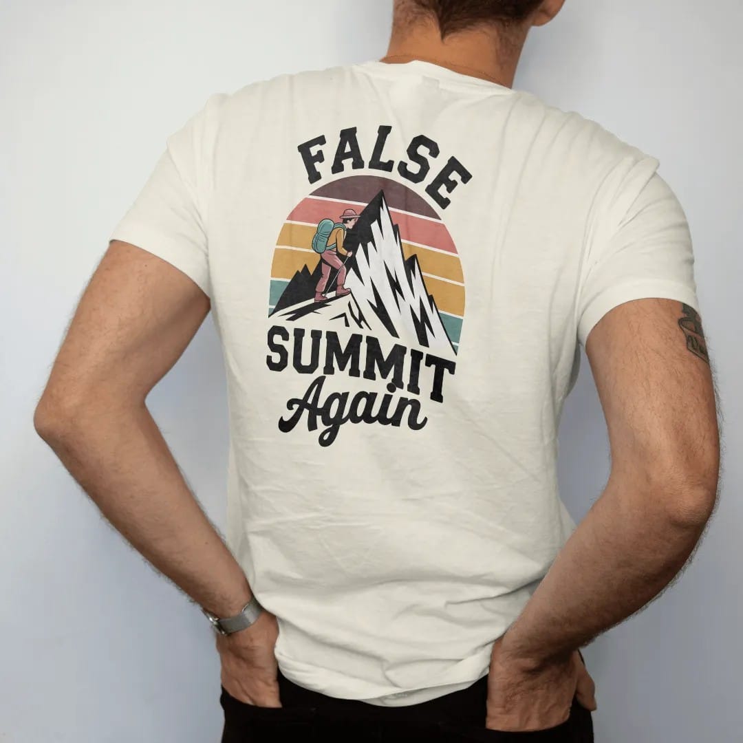 Man wearing a Ivory tee, seen from the back: a tired cartoon hiker cresting a ridge to see a taller peak behind it, stacked text above, faded 70s souvenir style, lettered "FALSE SUMMIT / AGAIN" in chunky rounded 70s letters with a drop shadow and a…
