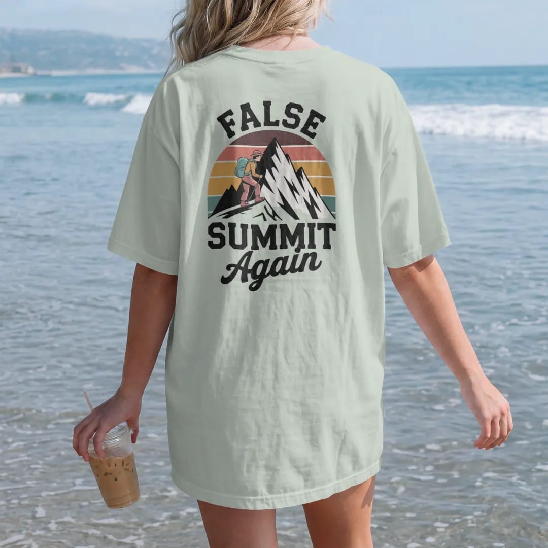 Woman on the beach in a Bay tee, back view: a tired cartoon hiker cresting a ridge to see a taller peak behind it, stacked text above, faded 70s souvenir style, lettered "FALSE SUMMIT / AGAIN" in chunky rounded 70s letters with a drop shadow and a…