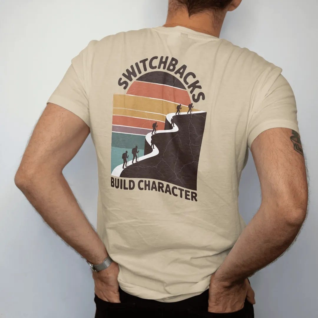 Man wearing a Sandstone tee, seen from the back: a zigzag trail climbing a steep mountain face drawn as a bold line with tiny hikers on each turn, faded 70s souvenir style, lettered "SWITCHBACKS / BUILD CHARACTER" in chunky rounded 70s letters with…