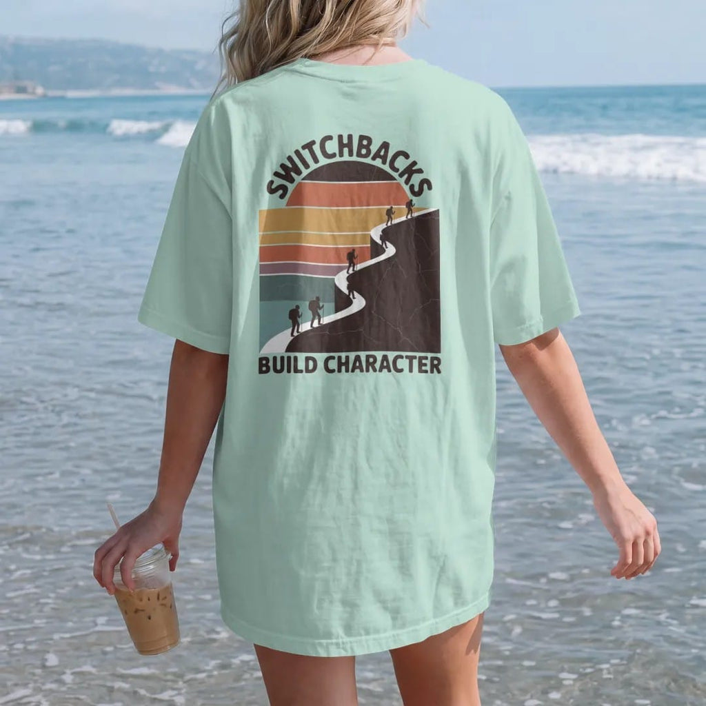 Woman on the beach in a Chalky Mint tee, back view: a zigzag trail climbing a steep mountain face drawn as a bold line with tiny hikers on each turn, faded 70s souvenir style, lettered "SWITCHBACKS / BUILD CHARACTER" in chunky rounded 70s letters…