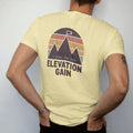 Man wearing a Butter tee, seen from the back: a bold elevation profile chart shaped like jagged peaks with a tiny top flag, faded 70s souvenir style, lettered "I CAME FOR THE / ELEVATION GAIN" in chunky rounded 70s letters with a drop shadow,…