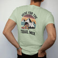 Man wearing a Light Green tee, seen from the back: a bulging zip bag of trail mix with peanuts, raisins and candy pieces spilling, mountains behind, faded 70s souvenir style, lettered "HERE FOR THE / TRAIL MIX" in chunky rounded 70s letters with a…