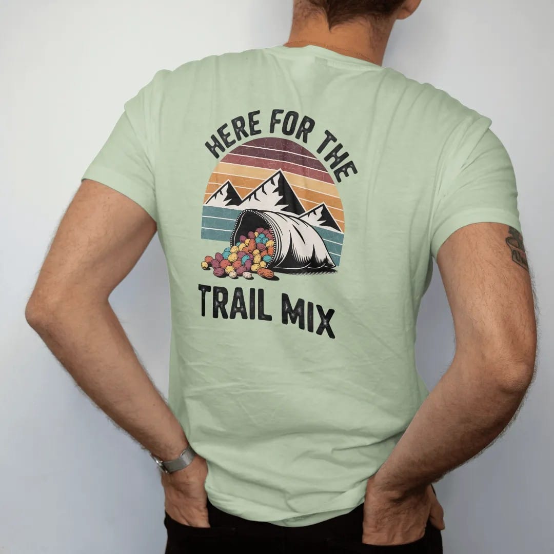 Man wearing a Light Green tee, seen from the back: a bulging zip bag of trail mix with peanuts, raisins and candy pieces spilling, mountains behind, faded 70s souvenir style, lettered "HERE FOR THE / TRAIL MIX" in chunky rounded 70s letters with a…