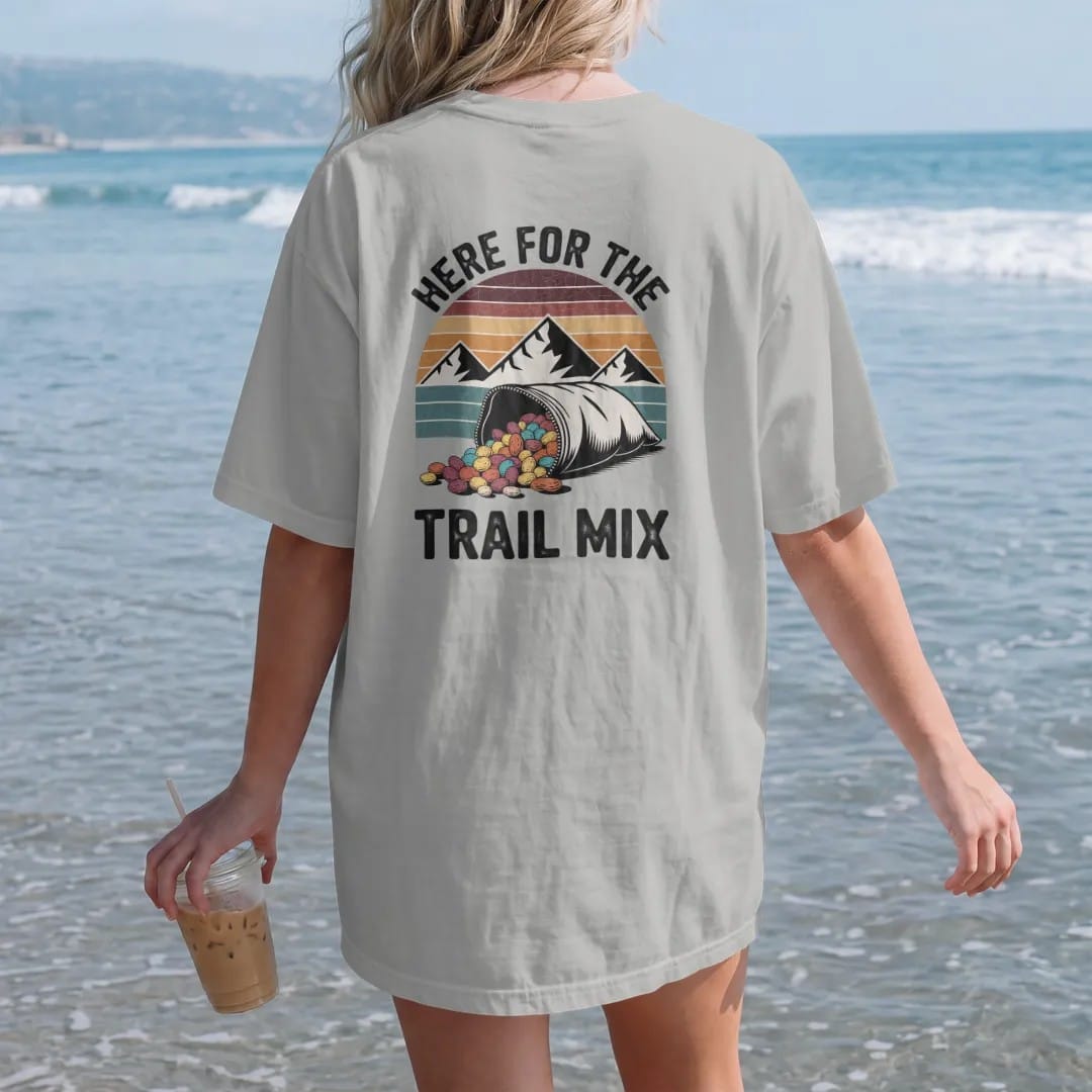 Woman on the beach in a Granite tee, back view: a bulging zip bag of trail mix with peanuts, raisins and candy pieces spilling, mountains behind, faded 70s souvenir style, lettered "HERE FOR THE / TRAIL MIX" in chunky rounded 70s letters with a…