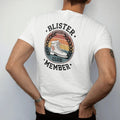 Man wearing a White tee, seen from the back: a battered hiking boot with a bandaged heel inside a round club emblem with laurels, faded 70s souvenir style, lettered "BLISTER CLUB / MEMBER" in chunky rounded 70s letters with a drop shadow, printed…