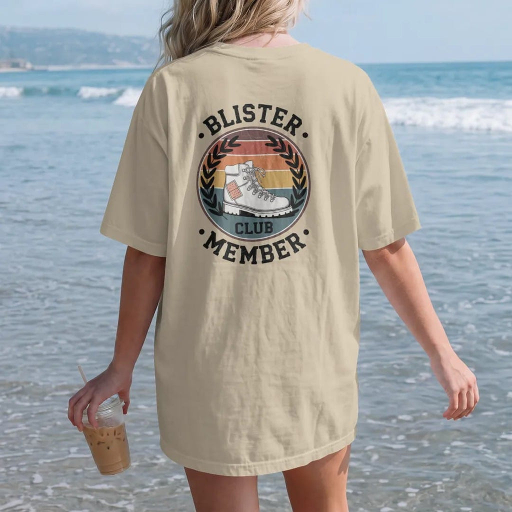 Woman on the beach in a Sandstone tee, back view: a battered hiking boot with a bandaged heel inside a round club emblem with laurels, faded 70s souvenir style, lettered "BLISTER CLUB / MEMBER" in chunky rounded 70s letters with a drop shadow,…