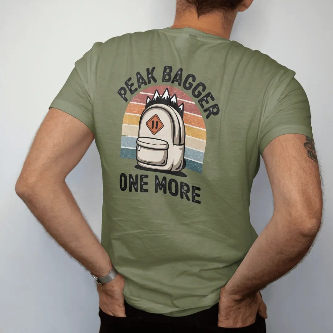 Man wearing a Moss tee, seen from the back: a cartoon backpack stuffed with tiny mountain peaks poking out of the top, shield badge, faded 70s souvenir style, lettered "PEAK BAGGER / ONE MORE" in chunky rounded 70s letters with a drop shadow,…