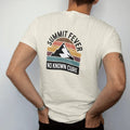 Man wearing a Ivory tee, seen from the back: a vintage medicine-label badge with a mountain peak and a thermometer reading off the chart, faded 70s souvenir style, lettered "SUMMIT FEVER / NO KNOWN CURE" in chunky rounded 70s serif letters with a…