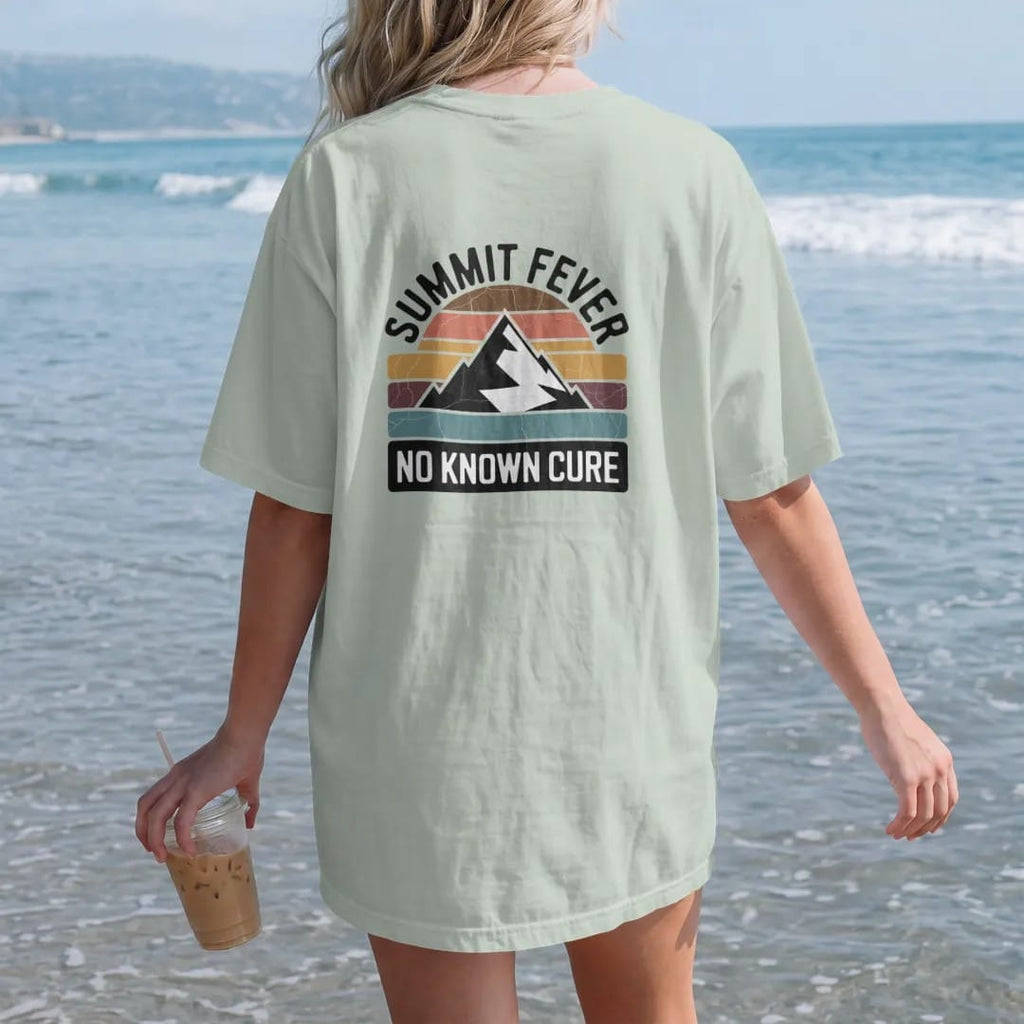 Woman on the beach in a Bay tee, back view: a vintage medicine-label badge with a mountain peak and a thermometer reading off the chart, faded 70s souvenir style, lettered "SUMMIT FEVER / NO KNOWN CURE" in chunky rounded 70s serif letters with a…