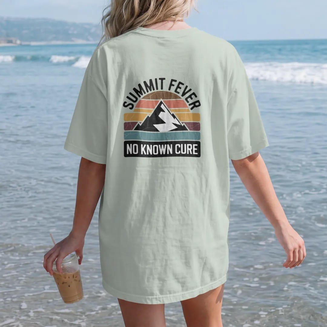 Woman on the beach in a Bay tee, back view: a vintage medicine-label badge with a mountain peak and a thermometer reading off the chart, faded 70s souvenir style, lettered "SUMMIT FEVER / NO KNOWN CURE" in chunky rounded 70s serif letters with a…