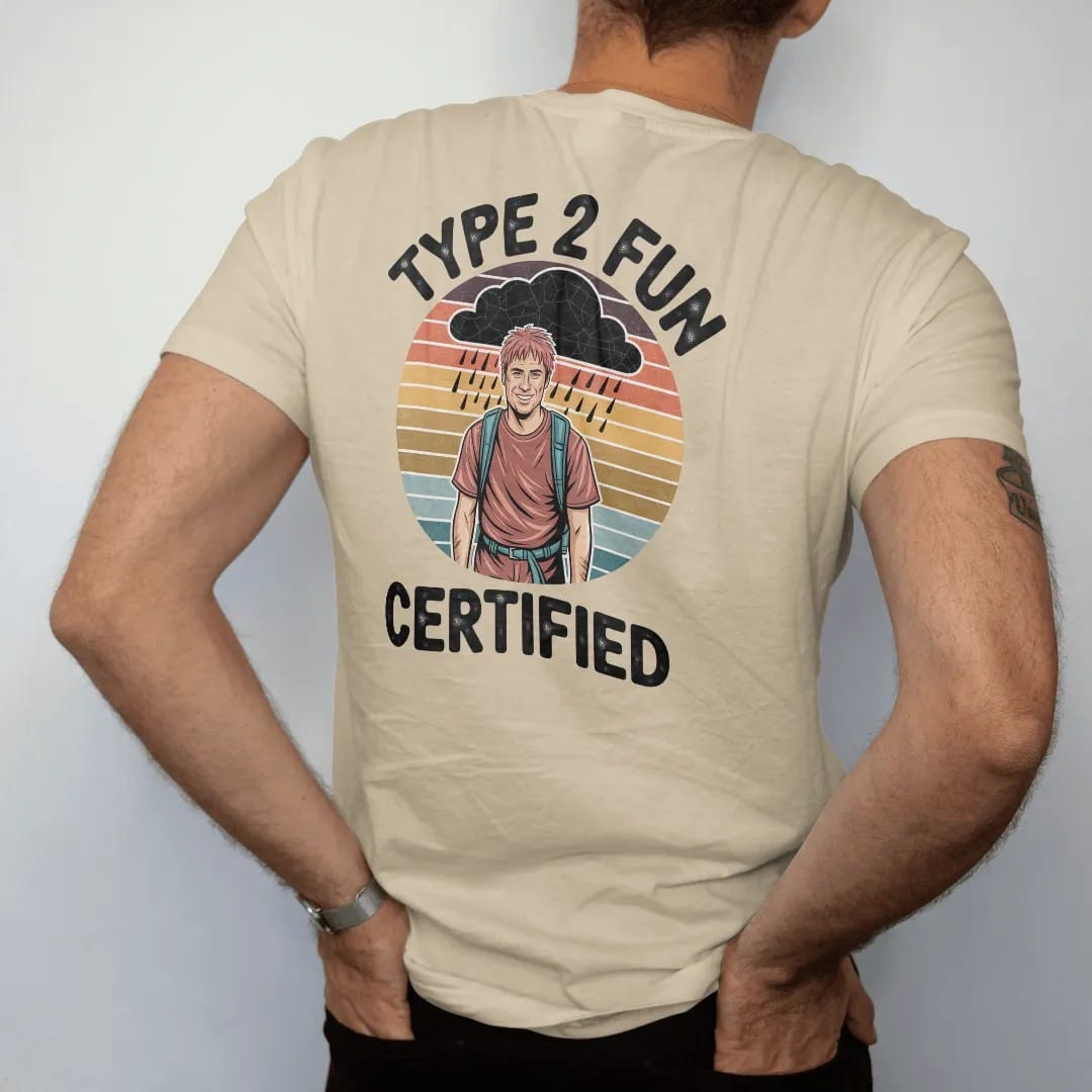 Man wearing a Sandstone tee, seen from the back: a muddy, rain-soaked hiker grinning under a storm cloud with a round certified seal, faded 70s souvenir style, lettered "TYPE 2 FUN / CERTIFIED" in chunky rounded 70s letters with a drop shadow,…