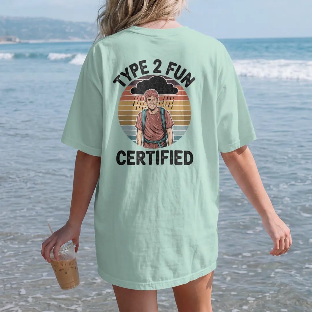 Woman on the beach in a Chalky Mint tee, back view: a muddy, rain-soaked hiker grinning under a storm cloud with a round certified seal, faded 70s souvenir style, lettered "TYPE 2 FUN / CERTIFIED" in chunky rounded 70s letters with a drop shadow,…