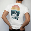 Man wearing a White tee, seen from the back: a single winding trail splitting into several paths through hills, each with a tiny hiker, faded 70s souvenir style, lettered "HIKE YOUR OWN HIKE" in chunky rounded 70s letters with a drop shadow,…