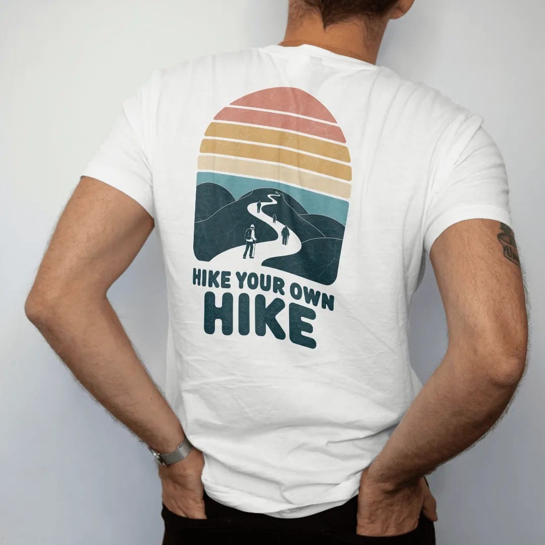 Man wearing a White tee, seen from the back: a single winding trail splitting into several paths through hills, each with a tiny hiker, faded 70s souvenir style, lettered "HIKE YOUR OWN HIKE" in chunky rounded 70s letters with a drop shadow,…