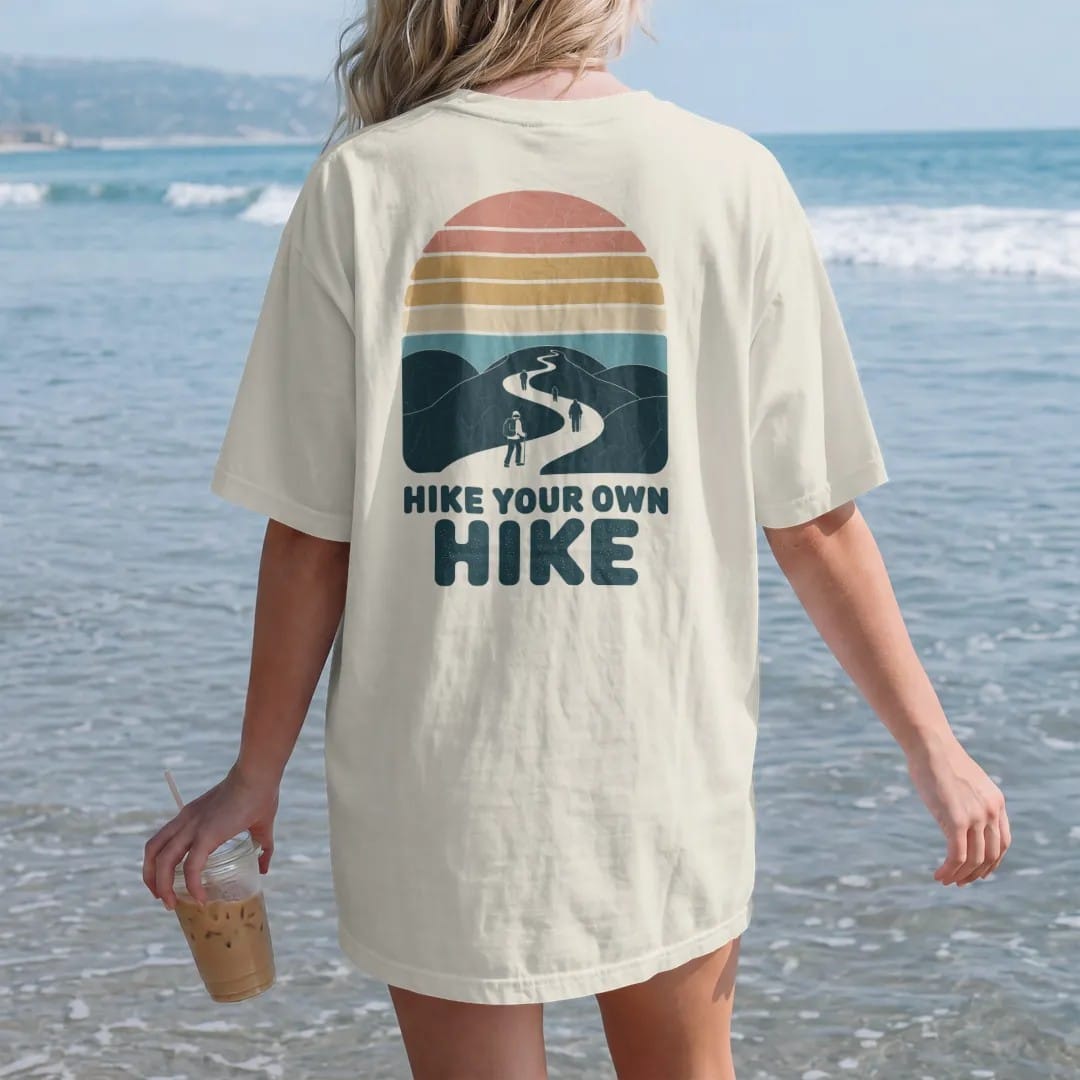 Woman on the beach in a Ivory tee, back view: a single winding trail splitting into several paths through hills, each with a tiny hiker, faded 70s souvenir style, lettered "HIKE YOUR OWN HIKE" in chunky rounded 70s letters with a drop shadow,…