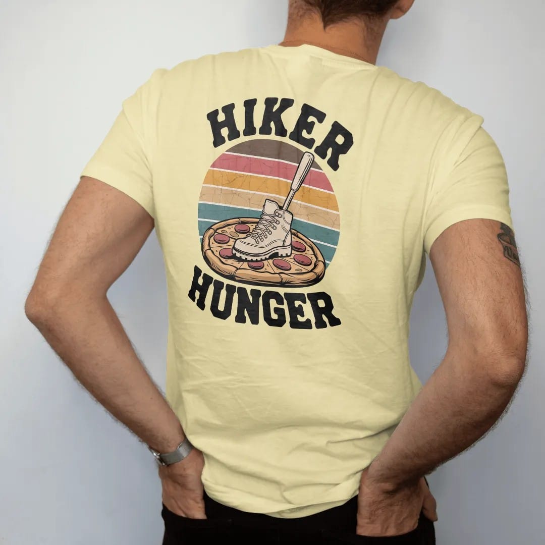 Man wearing a Butter tee, seen from the back: a whole pizza with a hiking boot print on it and a fork stuck in it like a summit flag, faded 70s souvenir style, lettered "HIKER HUNGER" in chunky rounded 70s letters with a drop shadow, printed in…