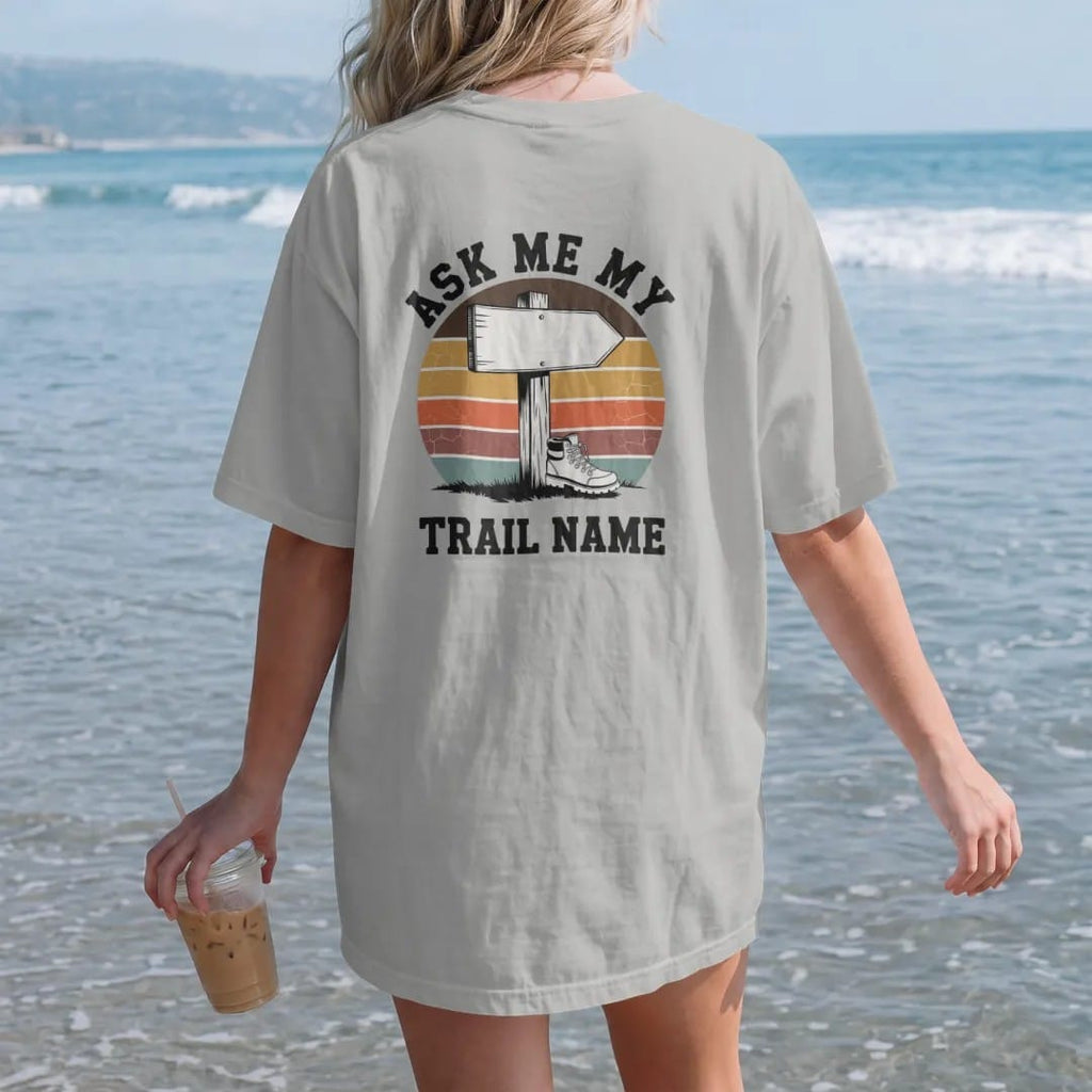 Woman on the beach in a Granite tee, back view: a wooden trail sign post with a blank arrow plank and a small hiking boot nailed to it, faded 70s souvenir style, lettered "ASK ME MY / TRAIL NAME" in chunky rounded 70s letters with a drop shadow,…
