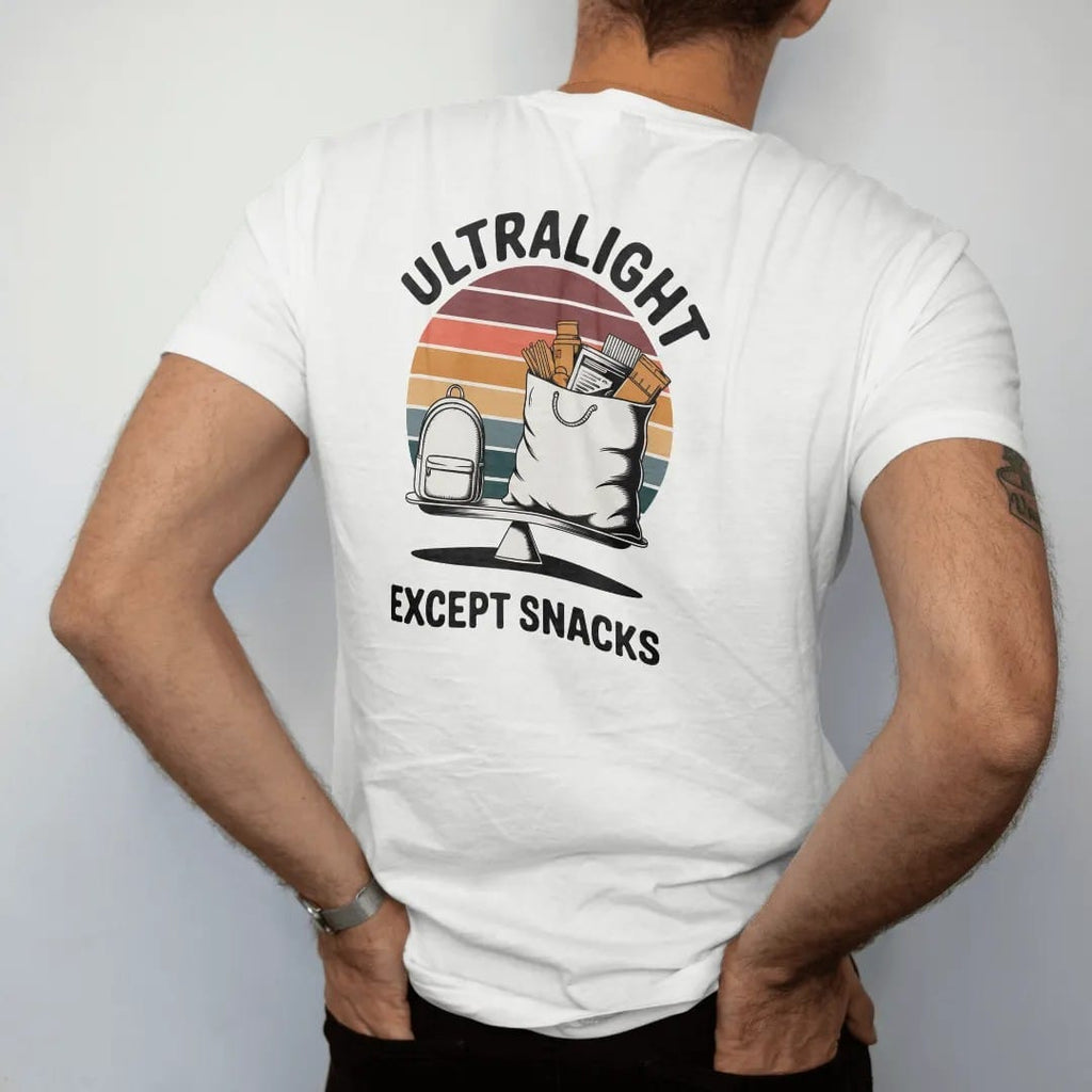 Man wearing a White tee, seen from the back: a tiny minimal backpack next to an enormous overflowing food bag on a scale, faded 70s souvenir style, lettered "ULTRALIGHT / EXCEPT SNACKS" in chunky rounded 70s letters with a drop shadow, printed in…