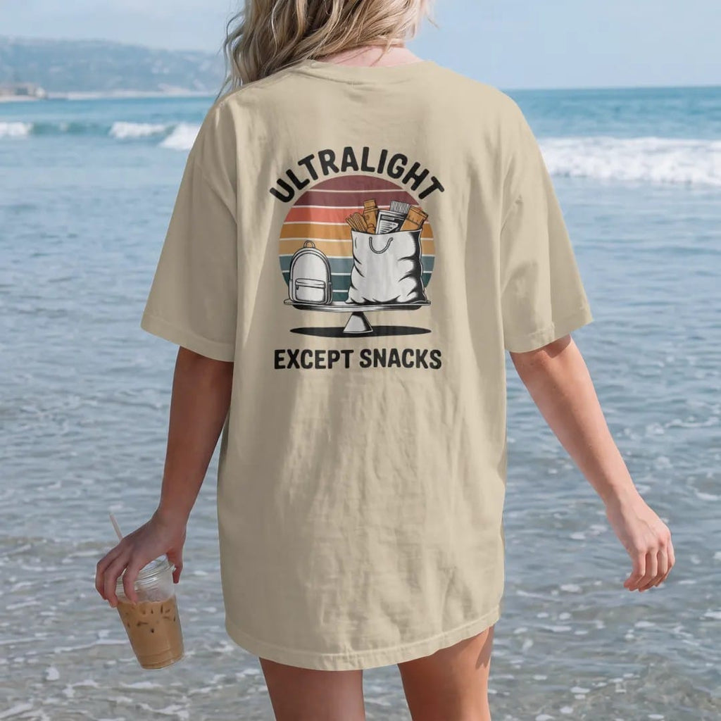 Woman on the beach in a Sandstone tee, back view: a tiny minimal backpack next to an enormous overflowing food bag on a scale, faded 70s souvenir style, lettered "ULTRALIGHT / EXCEPT SNACKS" in chunky rounded 70s letters with a drop shadow, printed…