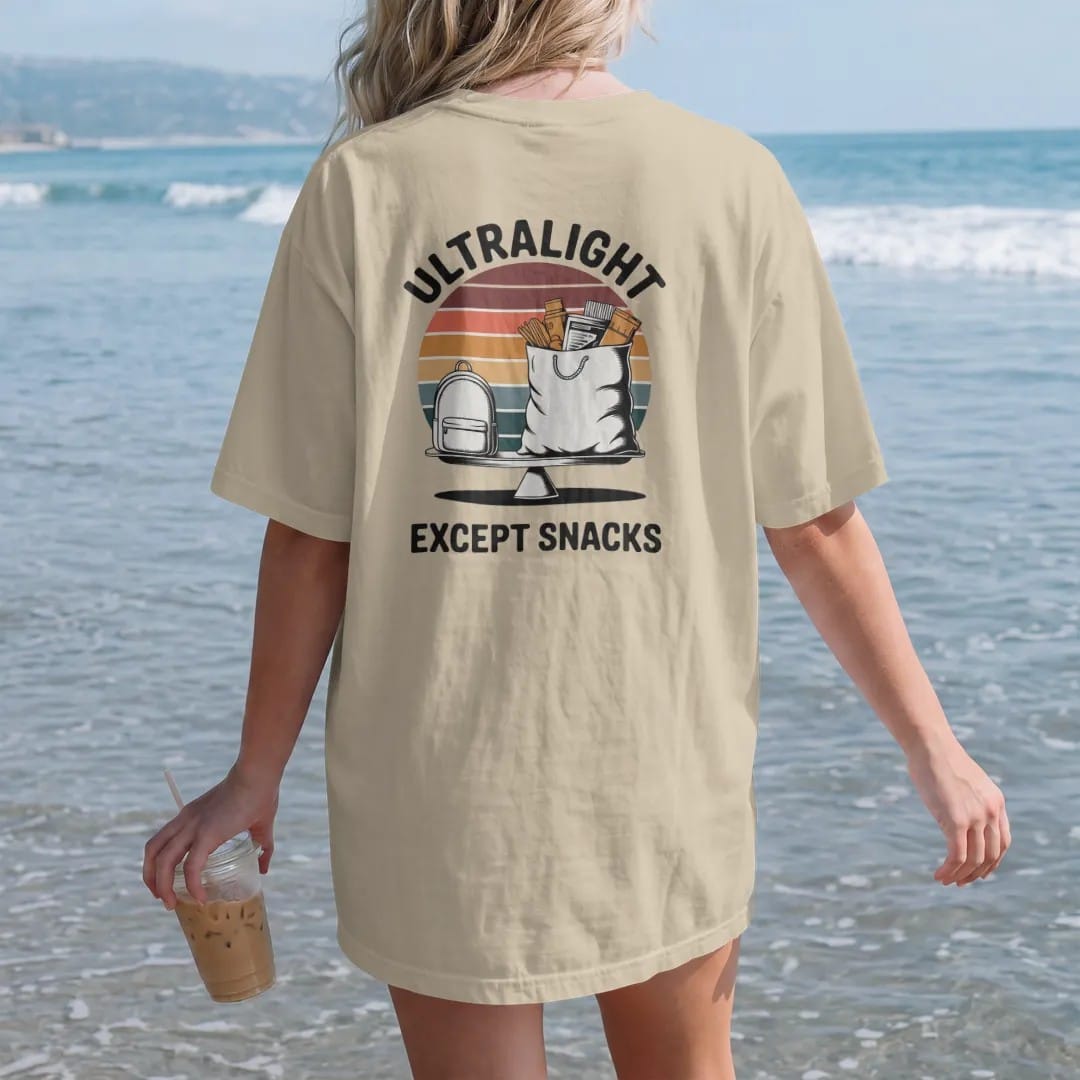 Woman on the beach in a Sandstone tee, back view: a tiny minimal backpack next to an enormous overflowing food bag on a scale, faded 70s souvenir style, lettered "ULTRALIGHT / EXCEPT SNACKS" in chunky rounded 70s letters with a drop shadow, printed…