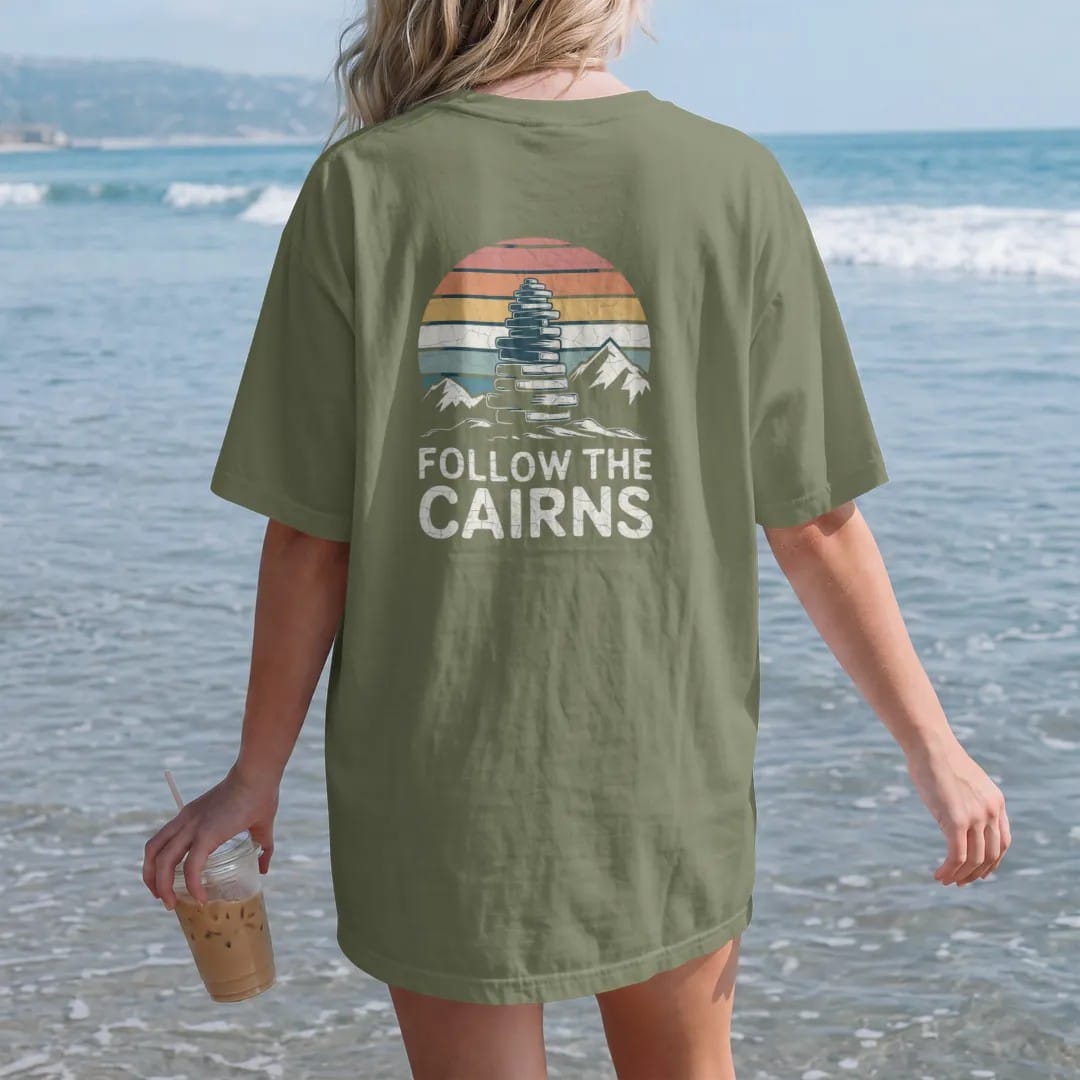 Woman on the beach in a Moss tee, back view: a stack of balanced trail cairns leading across an alpine slope toward a peak, faded 70s souvenir style, lettered "FOLLOW THE CAIRNS" in chunky rounded 70s letters with a drop shadow, printed in faded…