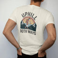 Man wearing a Ivory tee, seen from the back: a cartoon hiker leaning on trekking poles between two mountains that both slope upward, faded 70s souvenir style, lettered "UPHILL / BOTH WAYS" in chunky rounded 70s letters with a drop shadow, printed…
