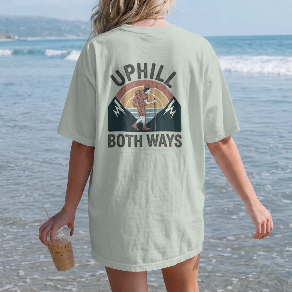 Woman on the beach in a Bay tee, back view: a cartoon hiker leaning on trekking poles between two mountains that both slope upward, faded 70s souvenir style, lettered "UPHILL / BOTH WAYS" in chunky rounded 70s letters with a drop shadow, printed in…