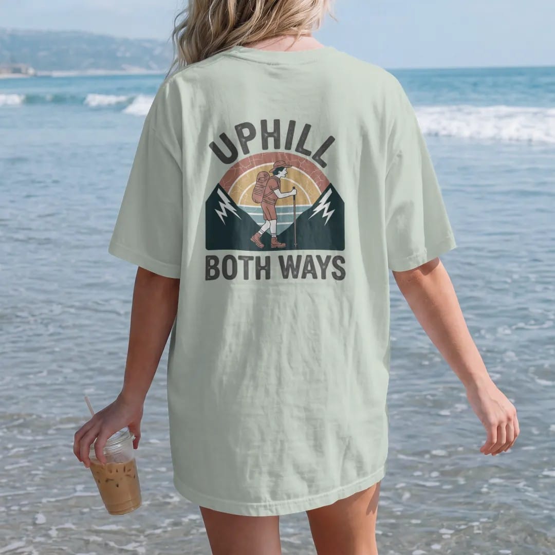 Woman on the beach in a Bay tee, back view: a cartoon hiker leaning on trekking poles between two mountains that both slope upward, faded 70s souvenir style, lettered "UPHILL / BOTH WAYS" in chunky rounded 70s letters with a drop shadow, printed in…