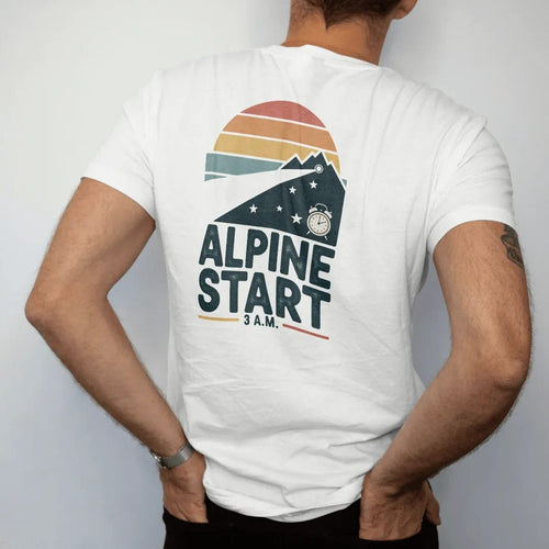 Man wearing a White tee, seen from the back: a headlamp beam cutting through the dark up a mountain with stars and a small alarm clock, faded 70s souvenir style, lettered "ALPINE START / 3 A.M." in chunky rounded 70s letters with a drop shadow,…
