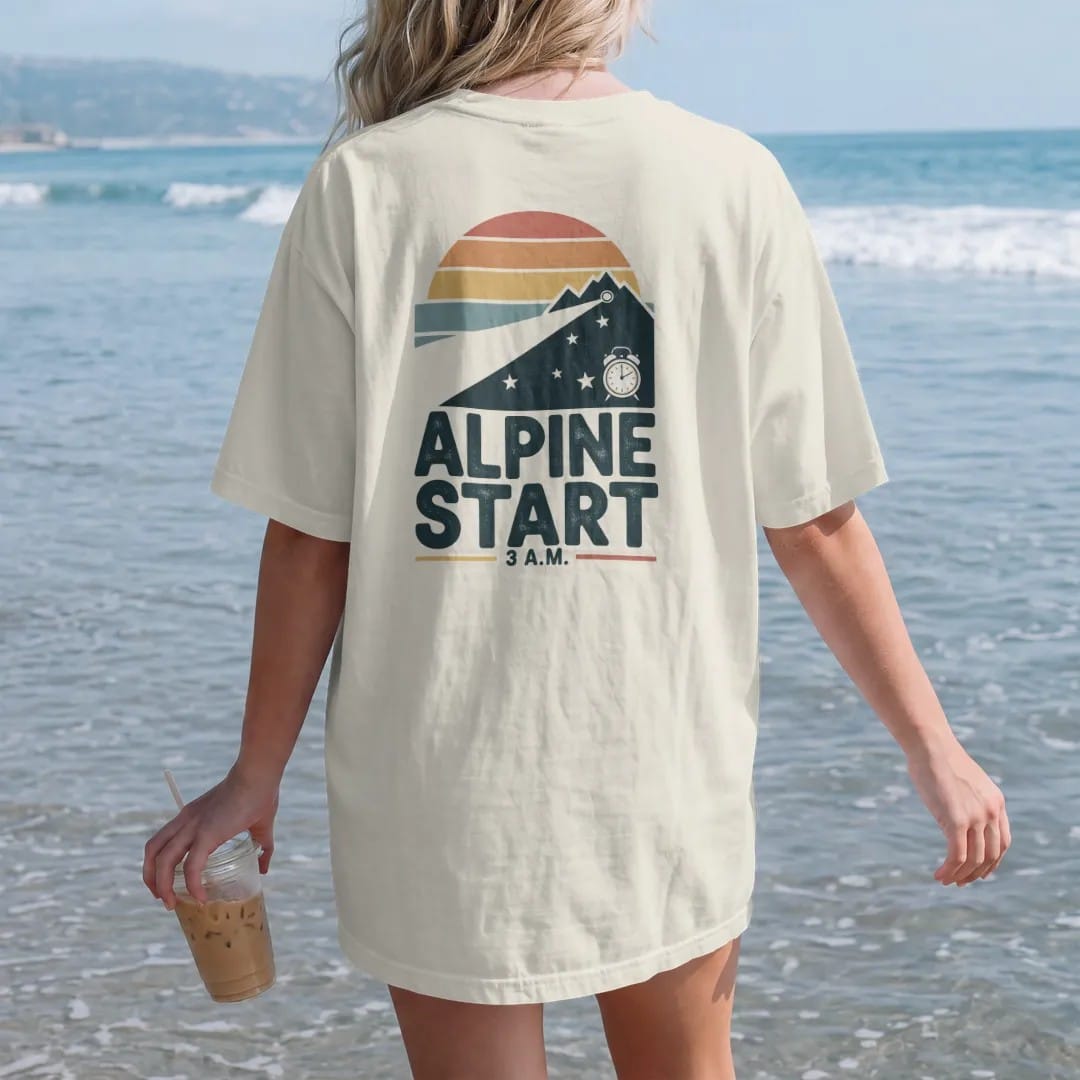 Woman on the beach in a Ivory tee, back view: a headlamp beam cutting through the dark up a mountain with stars and a small alarm clock, faded 70s souvenir style, lettered "ALPINE START / 3 A.M." in chunky rounded 70s letters with a drop shadow,…