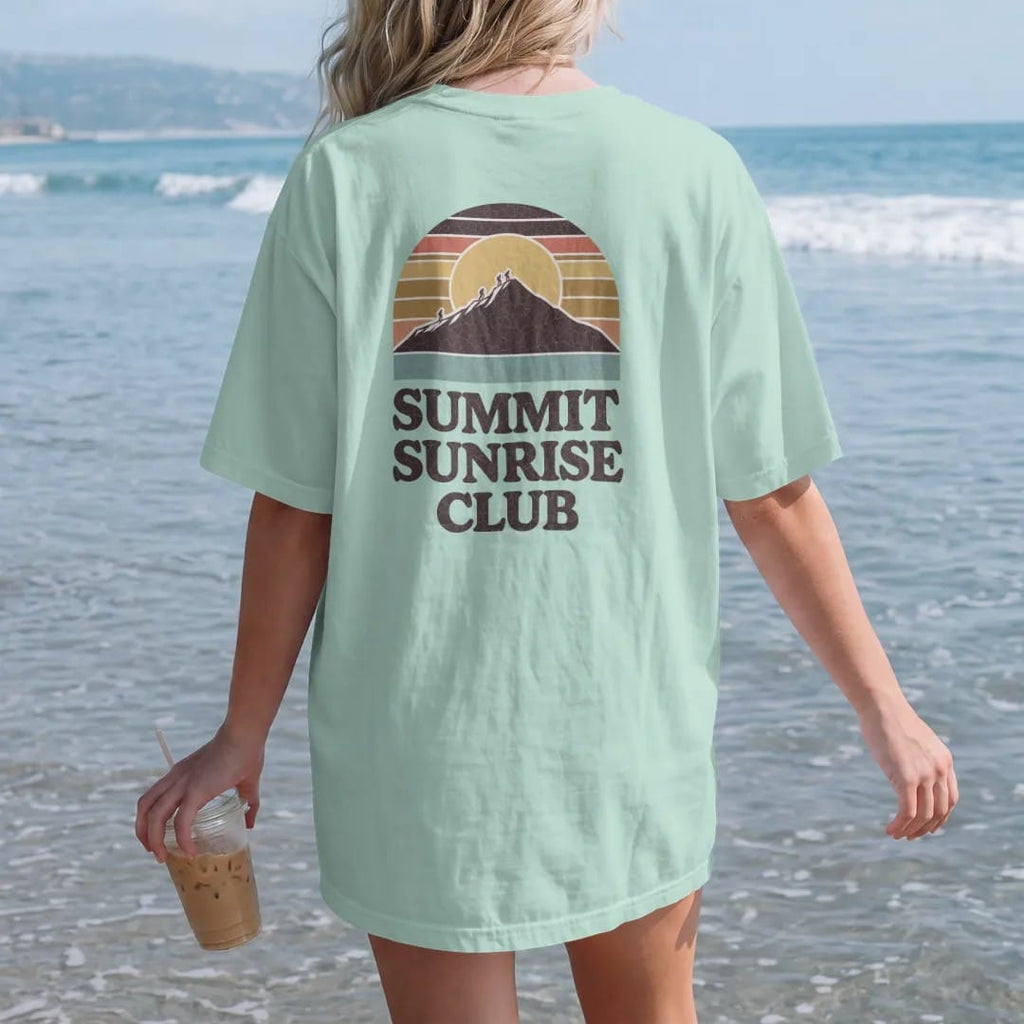 Woman on the beach in a Chalky Mint tee, back view: a huge rising sun behind a mountain peak with hikers silhouetted on top, round emblem, faded 70s souvenir style, lettered "SUMMIT SUNRISE CLUB" in chunky rounded 70s letters with a drop shadow,…