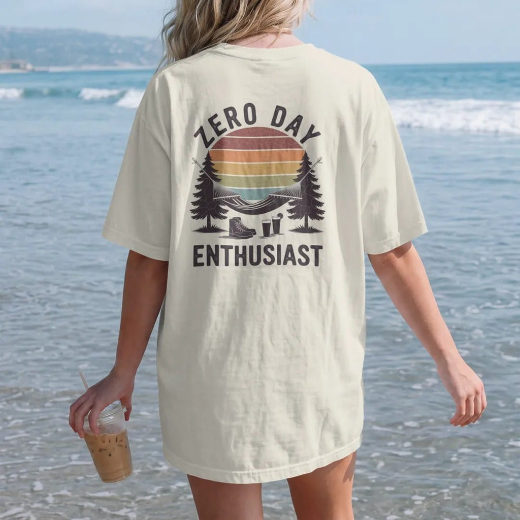 Woman on the beach in a Ivory tee, back view: a hammock strung between two pines with boots kicked off and a cold drink below, faded 70s souvenir style, lettered "ZERO DAY / ENTHUSIAST" in chunky rounded 70s letters with a drop shadow, printed in…