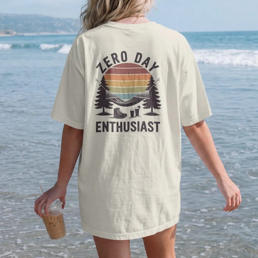 Woman on the beach in a Ivory tee, back view: a hammock strung between two pines with boots kicked off and a cold drink below, faded 70s souvenir style, lettered "ZERO DAY / ENTHUSIAST" in chunky rounded 70s letters with a drop shadow, printed in…