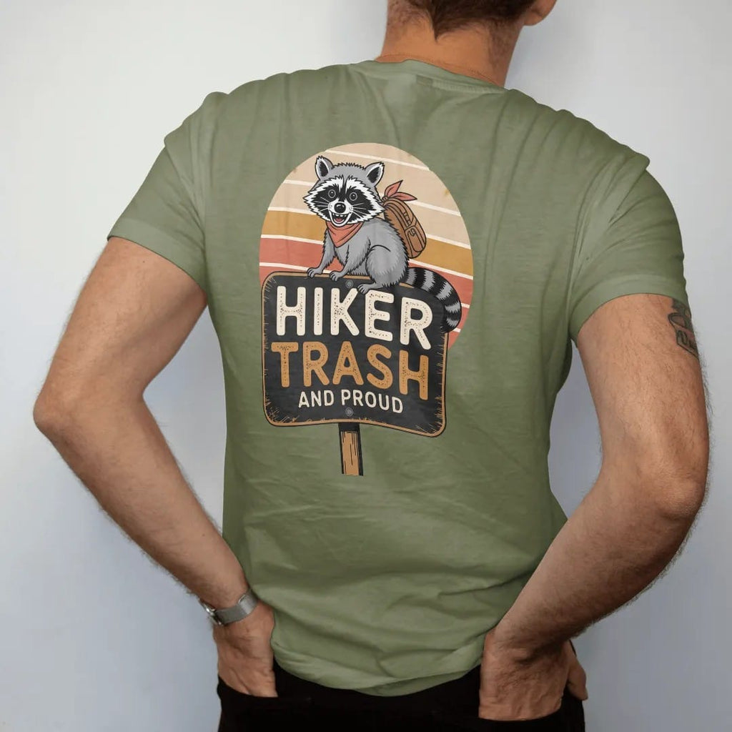 Man wearing a Moss tee, seen from the back: a scruffy grinning raccoon wearing a bandana and a tiny backpack on a trail sign, faded 70s souvenir style, lettered "HIKER TRASH / AND PROUD" in chunky rounded 70s letters with a drop shadow, printed in…