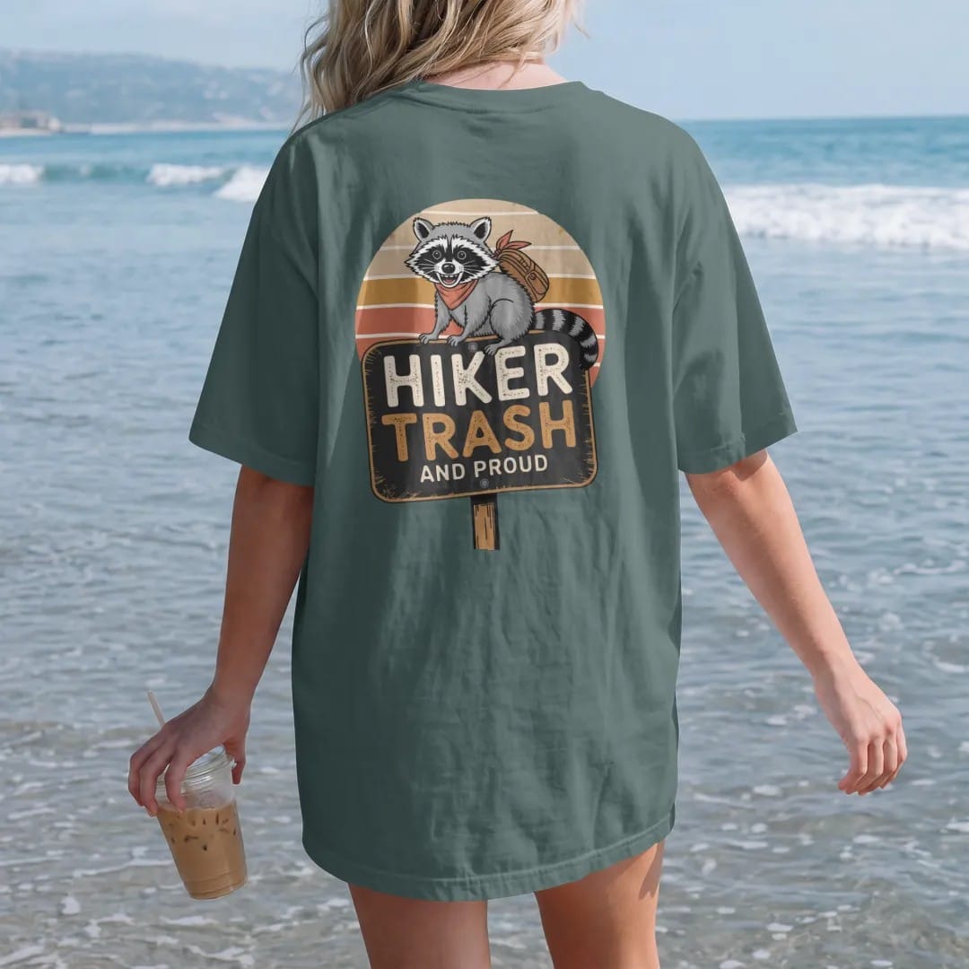 Woman on the beach in a Blue Spruce tee, back view: a scruffy grinning raccoon wearing a bandana and a tiny backpack on a trail sign, faded 70s souvenir style, lettered "HIKER TRASH / AND PROUD" in chunky rounded 70s letters with a drop shadow,…