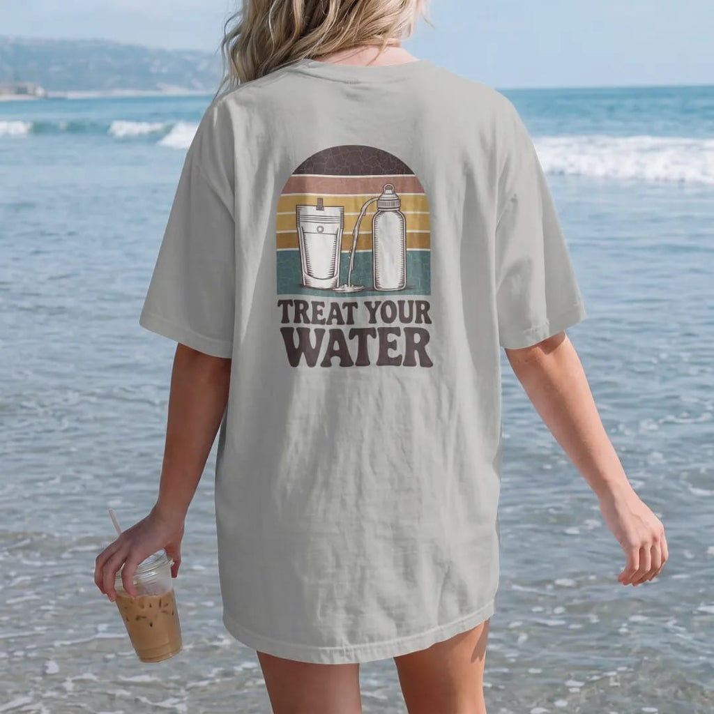 Woman on the beach in a Granite tee, back view: a mountain stream flowing into a squeeze filter pouch and a water bottle, field-guide layout, faded 70s souvenir style, lettered "TREAT YOUR WATER" in chunky rounded 70s serif letters with a drop…