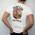 Man wearing a White tee, seen from the back: an open bear canister with food items stacked in tidy blocks and a bear peeking from behind a rock, faded 70s souvenir style, lettered "BEAR CAN / PUZZLE MASTER" in chunky rounded 70s letters with a drop…