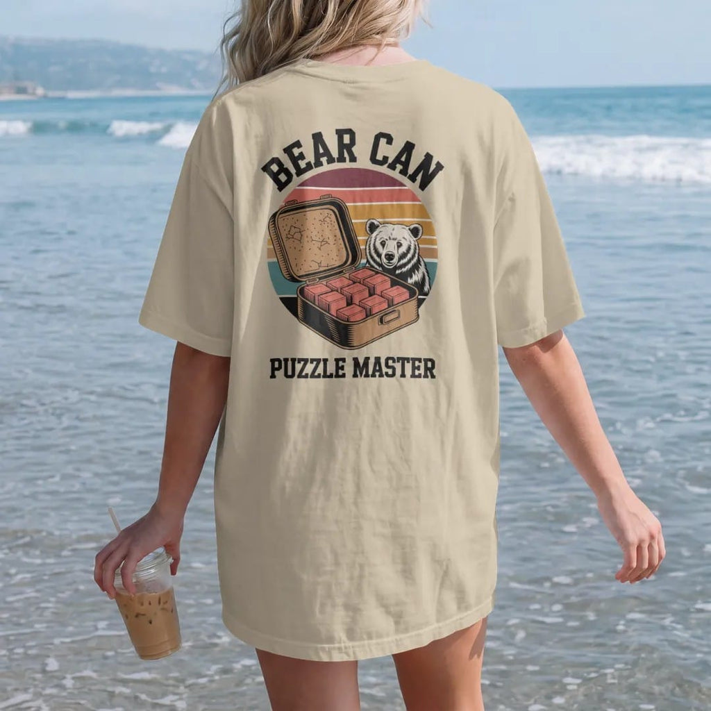 Woman on the beach in a Sandstone tee, back view: an open bear canister with food items stacked in tidy blocks and a bear peeking from behind a rock, faded 70s souvenir style, lettered "BEAR CAN / PUZZLE MASTER" in chunky rounded 70s letters with a…