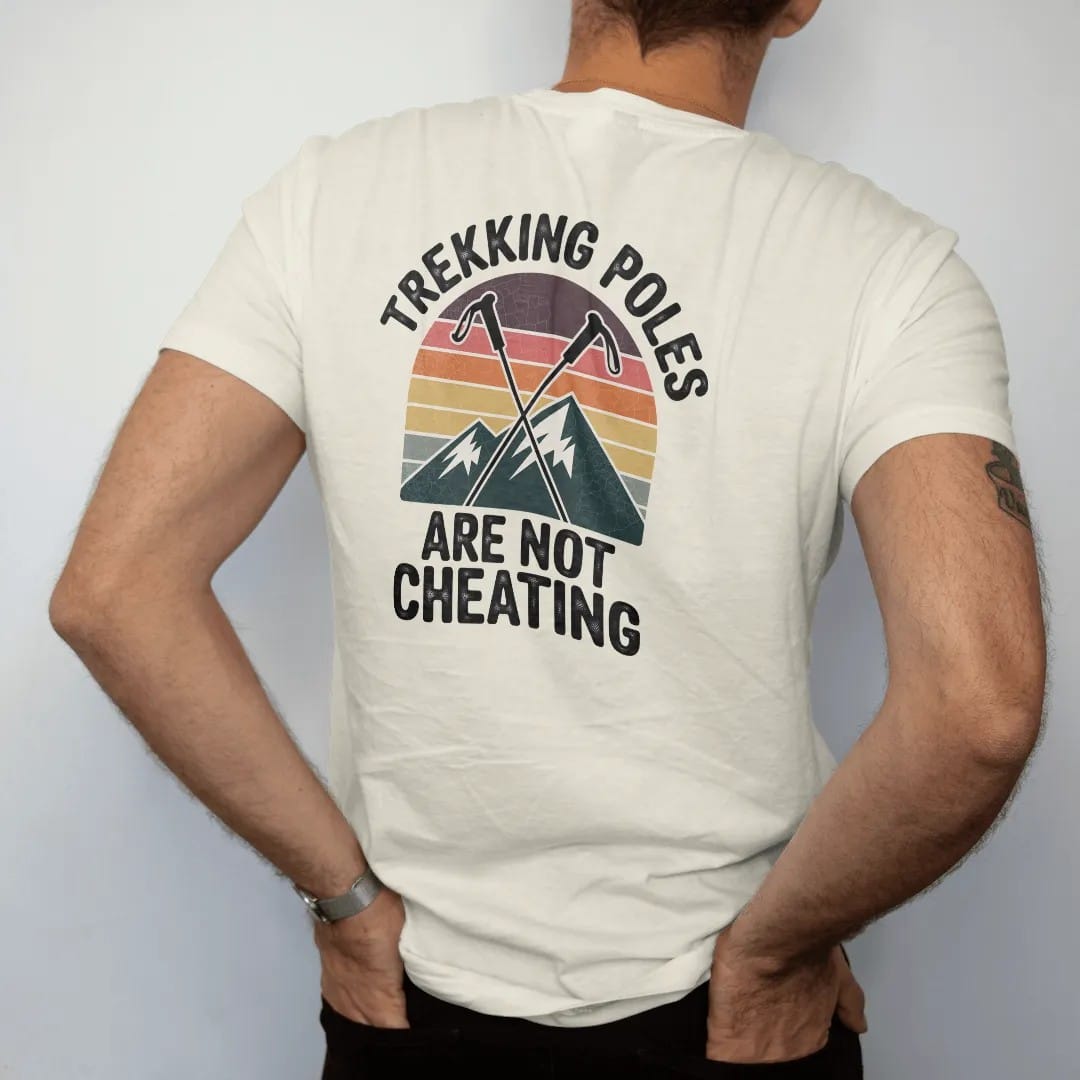 Man wearing a Ivory tee, seen from the back: crossed trekking poles over a steep mountain with a pair of happy cartoon knees, faded 70s souvenir style, lettered "TREKKING POLES / ARE NOT CHEATING" in chunky rounded 70s letters with a drop shadow,…