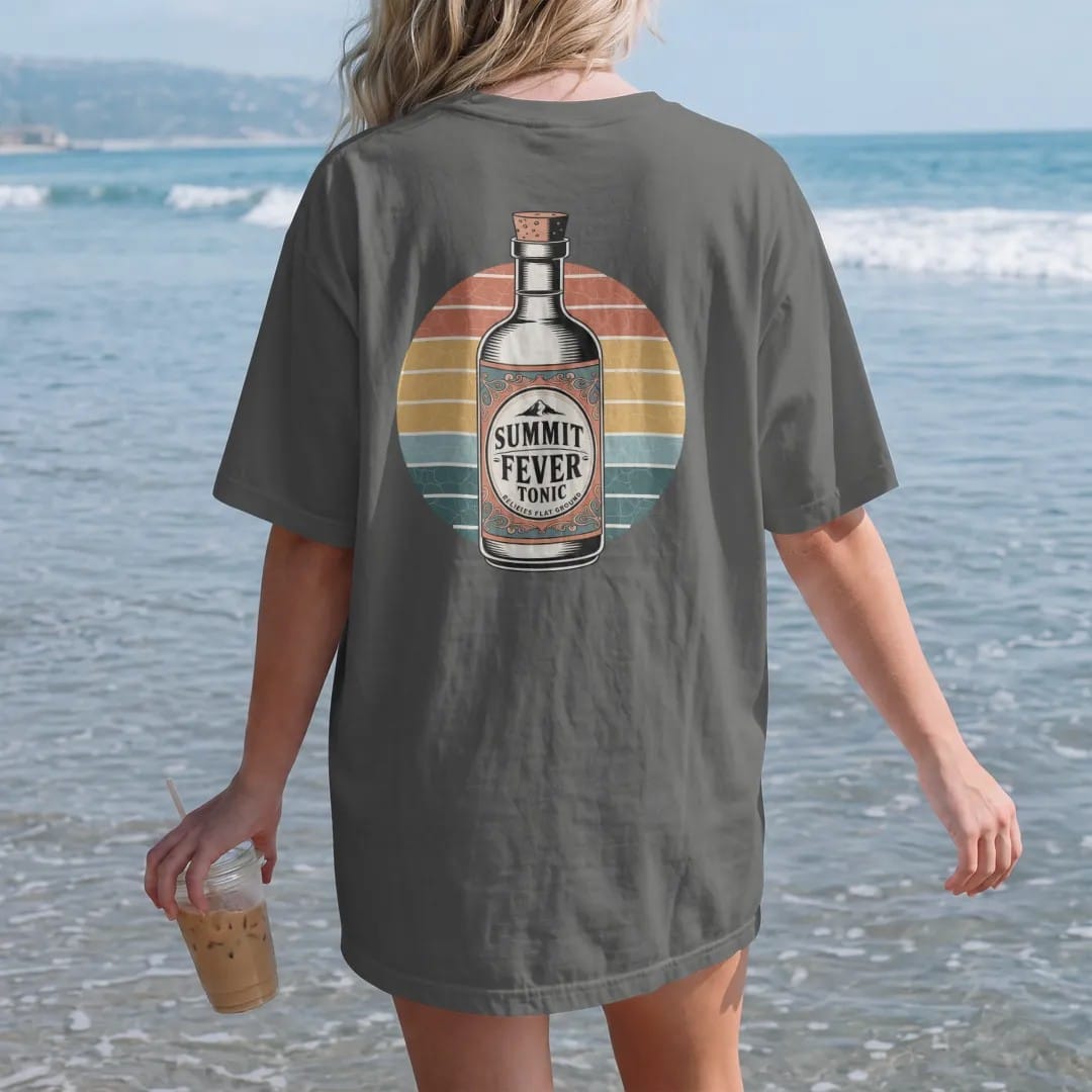 Woman on the beach in a Pepper tee, back view: an antique tonic bottle with a mountain on the label, ornate scrolls and tiny banners, faded 70s souvenir style, lettered "SUMMIT FEVER TONIC / RELIEVES FLAT GROUND" in chunky rounded 70s serif letters…