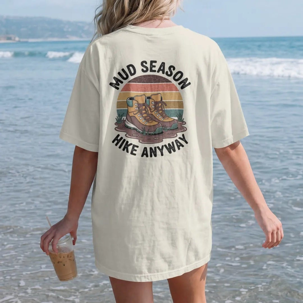 Woman on the beach in a Ivory tee, back view: a pair of hiking boots sunk deep in a mud puddle with spring sprouts and a few raindrops, faded 70s souvenir style, lettered "MUD SEASON / HIKE ANYWAY" in chunky rounded 70s letters with a drop shadow,…