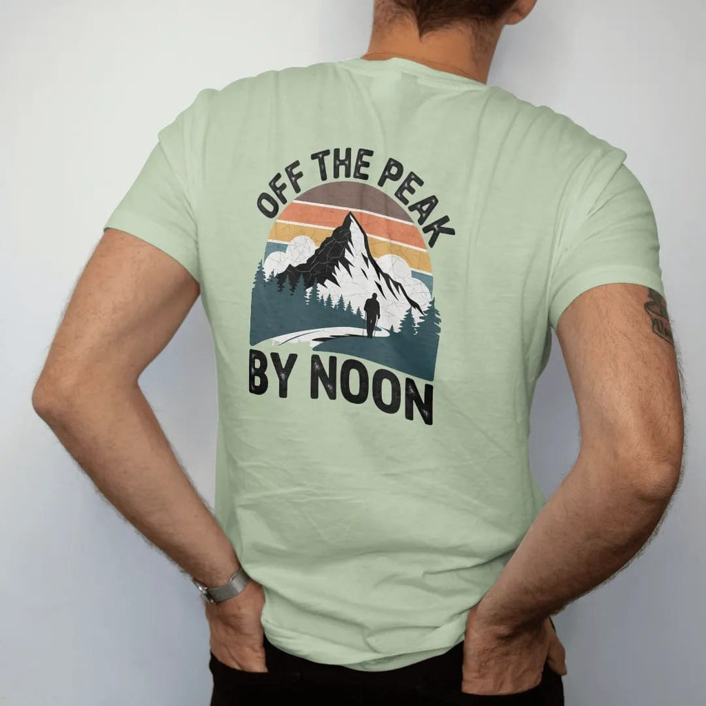 Man wearing a Light Green tee, seen from the back: a bright summer peak with puffy thunderheads building behind it and a hiker heading down, faded 70s souvenir style, lettered "OFF THE PEAK / BY NOON" in chunky rounded 70s letters with a drop…