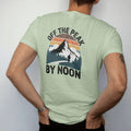 Man wearing a Light Green tee, seen from the back: a bright summer peak with puffy thunderheads building behind it and a hiker heading down, faded 70s souvenir style, lettered "OFF THE PEAK / BY NOON" in chunky rounded 70s letters with a drop…