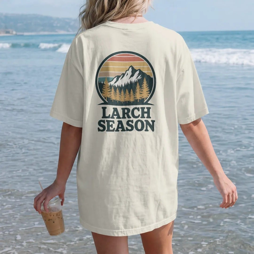 Woman on the beach in a Ivory tee, back view: golden larch trees glowing on a mountainside below a granite peak, round badge frame, faded 70s souvenir style, lettered "LARCH SEASON" in chunky rounded 70s letters with a drop shadow, printed in faded…