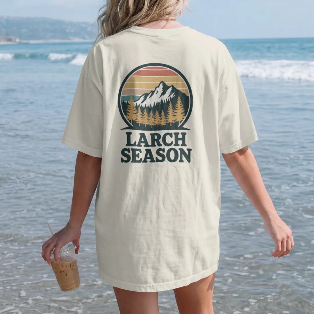 Woman on the beach in a Ivory tee, back view: golden larch trees glowing on a mountainside below a granite peak, round badge frame, faded 70s souvenir style, lettered "LARCH SEASON" in chunky rounded 70s letters with a drop shadow, printed in faded…