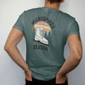 Man wearing a Blue Spruce tee, seen from the back: a boot wearing chain traction spikes on an icy trail with snowy pines and snowflakes, faded 70s souvenir style, lettered "MICROSPIKES / SEASON" in chunky rounded 70s letters with a drop shadow,…