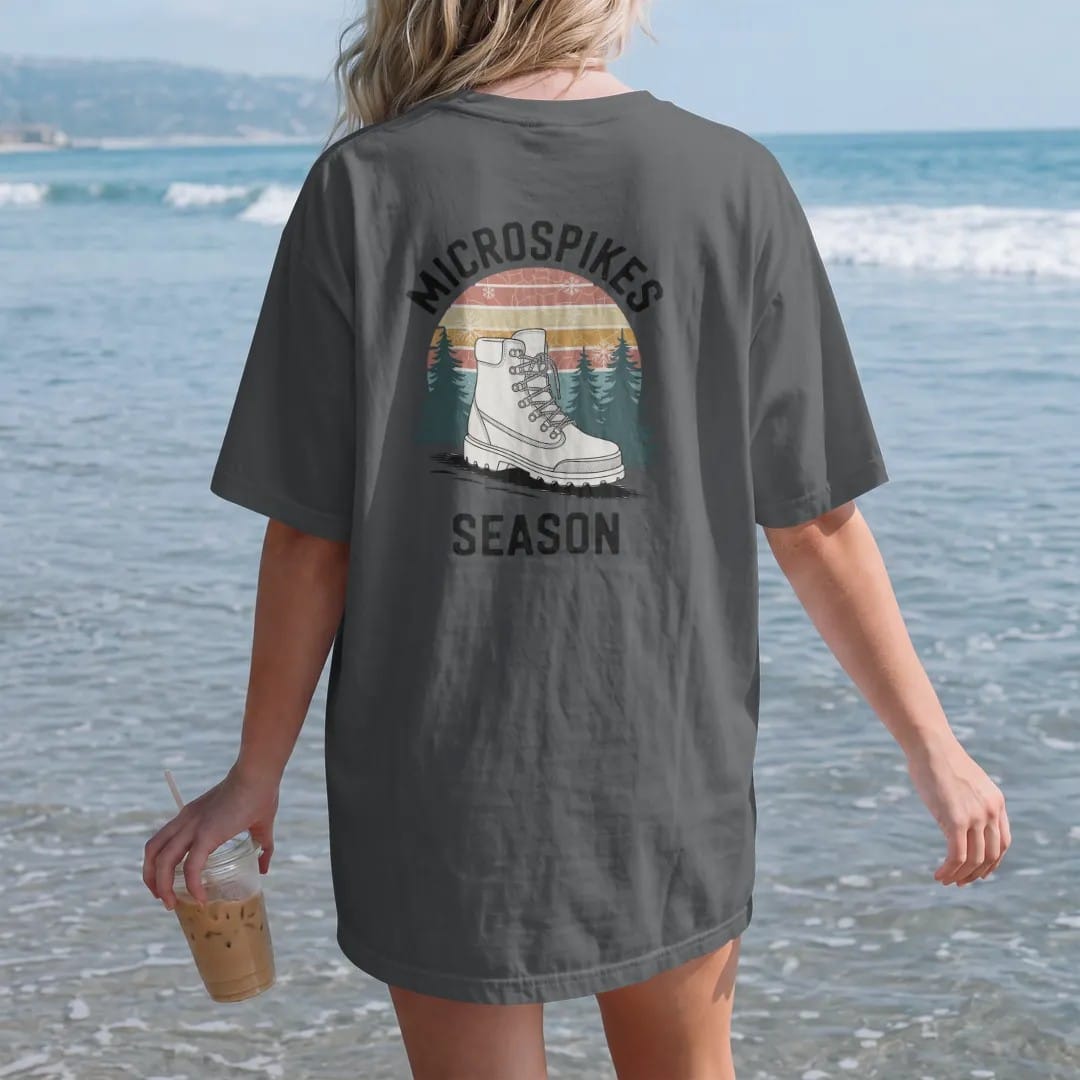 Woman on the beach in a Graphite tee, back view: a boot wearing chain traction spikes on an icy trail with snowy pines and snowflakes, faded 70s souvenir style, lettered "MICROSPIKES / SEASON" in chunky rounded 70s letters with a drop shadow,…